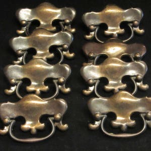 May include: Eight antique brass cabinet drawer pulls with ornate designs. The pulls have a loop handle and are in a distressed finish.