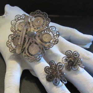 May include: A vintage silver filigree jewelry set featuring a large flower brooch and matching flower earrings. The brooch has a pin closure. The earrings are screw-back style. The set is displayed on a white surface.
