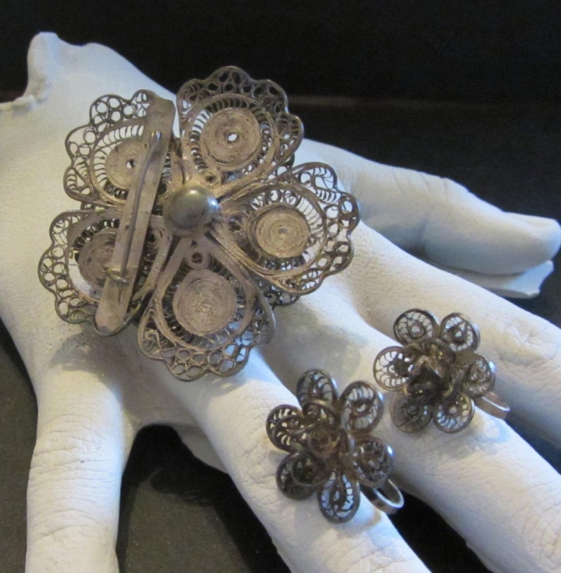 May include: A vintage silver filigree jewelry set featuring a large flower brooch and matching flower earrings. The brooch has a pin closure. The earrings are screw-back style. The set is displayed on a white surface.