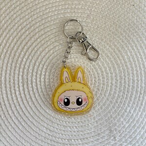 May include: A yellow acrylic keychain featuring a cartoon character in a bunny costume with a mischievous grin. The keychain has a silver keyring, chain, and clasp. The bunny's face has pink cheeks and black eyes.