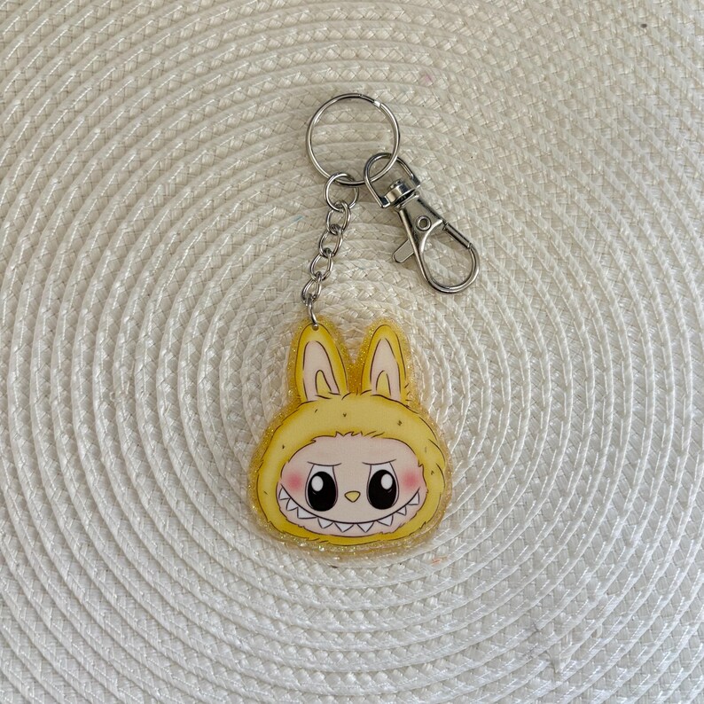May include: A yellow acrylic keychain featuring a cartoon character in a bunny costume with a mischievous grin. The keychain has a silver keyring, chain, and clasp. The bunny's face has pink cheeks and black eyes.