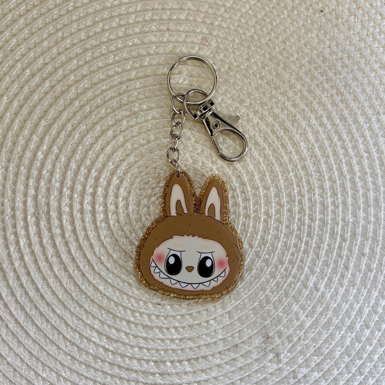 May include: A brown bunny-shaped keychain with a cartoon face and a gold glitter border. The keychain has a silver keyring and clasp. The bunny has white eyes, a pink blush, and a jagged-tooth grin. The keychain is on a white woven surface.