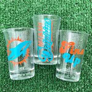 May include: Three clear shot glasses with different designs. One glass features the Miami Dolphins logo in orange and teal. Another says "Miami Dolphins" in teal and orange. The third says "Fins Up" in orange and teal.