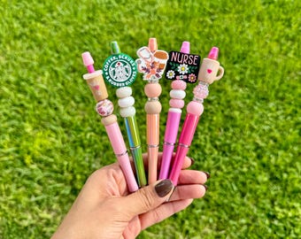 May include: Five pink and green pens with different charms. The charms include a coffee cup, a nurse's cap, a flower, and a Starbucks logo. The pens are decorated with beads.