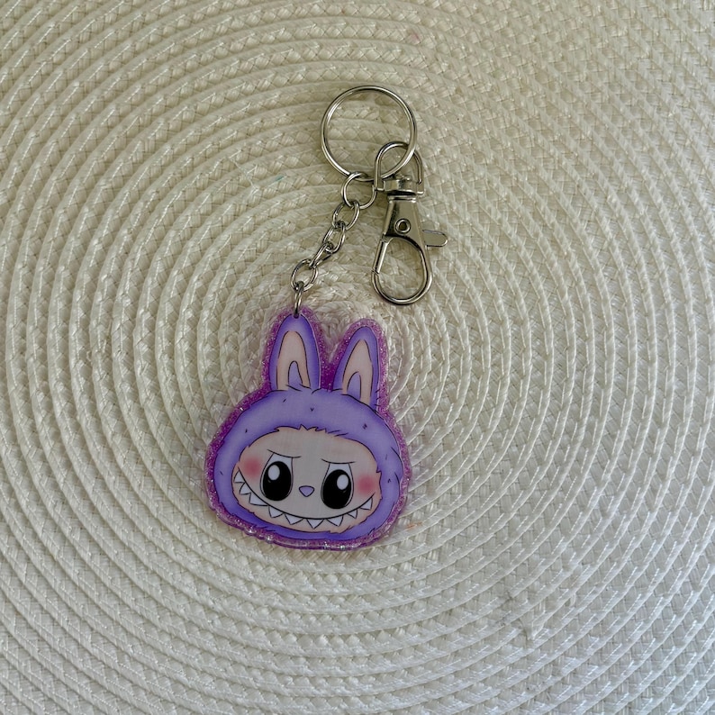 May include: A purple bunny-shaped keychain with a cartoon face and a silver keyring. The bunny has a mischievous grin and is wearing a hooded outfit. The keychain is made of acrylic and has a glittery border.