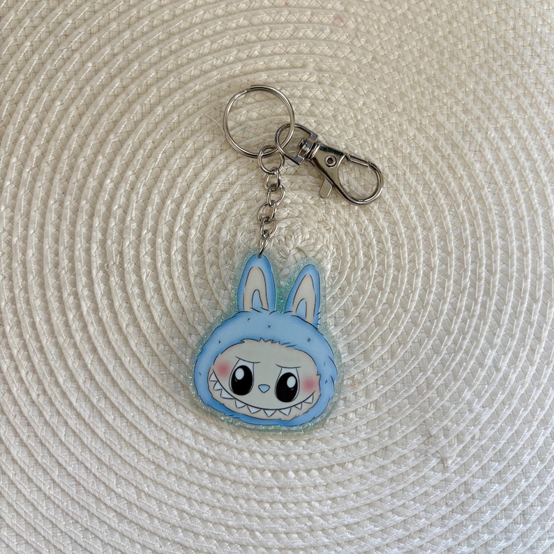 May include: A light blue acrylic keychain featuring a cartoon character in a bunny costume with a toothy grin. The keychain has a silver metal ring and clasp for attaching to keys or bags. The bunny's face has black eyes and pink cheeks.