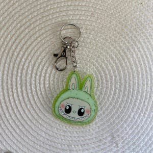 May include: A green and white bunny-themed keychain with a silver clasp and chain. The bunny has a mischievous expression with large black eyes and a toothy grin. The keychain is made of acrylic and has a glittery border.