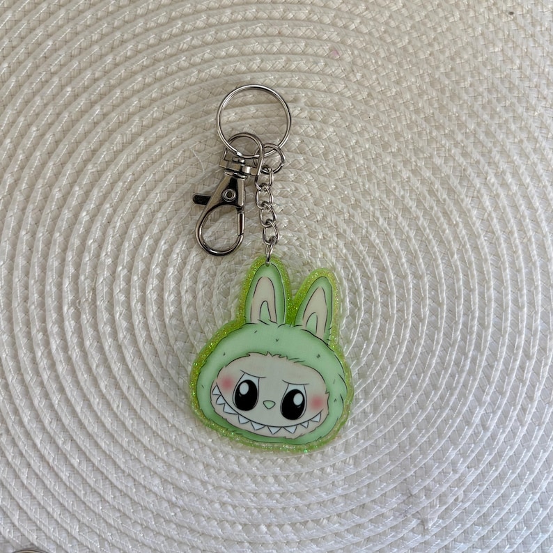 May include: A green and white bunny-themed keychain with a silver clasp and chain. The bunny has a mischievous expression with large black eyes and a toothy grin. The keychain is made of acrylic and has a glittery border.