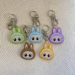 May include: Six colorful acrylic keychains featuring a cartoon bunny design with a toothy grin. The keychains are in shades of green, blue, brown, yellow, and purple, each with a silver keyring and clasp.