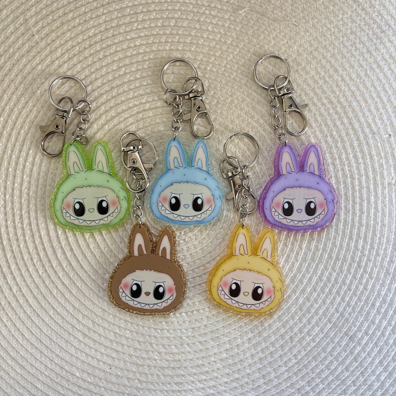 May include: Six colorful acrylic keychains featuring a cartoon bunny design with a toothy grin. The keychains are in shades of green, blue, brown, yellow, and purple, each with a silver keyring and clasp.
