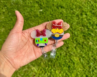 Puede incluir: Dos portatarjetas retráctiles con temática de personajes. Uno presenta un diseño verde, morado y blanco con un lazo rojo. El otro tiene un diseño de purpurina dorada con un lazo rojo y un cinturón azul y amarillo. Ambos tienen un clip de plástico transparente.