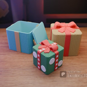 Może przedstawiać: Trzy pudełka prezentowe z dzianinowym wzorem. Jedno pudełko jest turkusowe z żółtym paskiem, drugie jest beżowe z czerwonym paskiem, a ostatnie jest zielone z czerwoną kokardką i białymi kropkami.