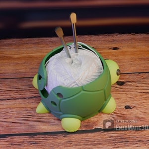 Puede incluir: Un cuenco de hilo verde con forma de tortuga que contiene una bola de lana blanca. Dos agujas de tejer plateadas están insertadas en la lana. El cuenco tiene patas amarillas y un diseño de caparazón texturizado.