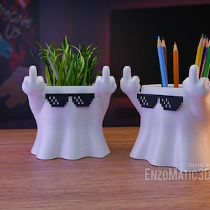 May include: Two white ghost-shaped planters with black pixelated sunglasses. One planter has a small green plant growing out of the top, and the other planter has a group of coloured pencils inside.