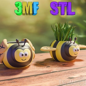 Puede incluir: Dos macetas en forma de abeja de color amarillo y negro con una linda cara de dibujos animados. Una maceta tiene hierba verde creciendo fuera de ella. Las macetas están etiquetadas como "3MF STL".