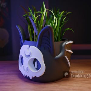 Puede incluir: Una maceta de cerámica negra y blanca con forma de murciélago con una cara de calavera y una luna creciente en la frente. La maceta está llena de hierba verde.