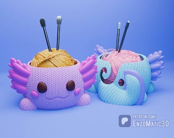 May include: Two axolotl-shaped yarn bowls, one purple and one blue, each holding yarn and knitting needles. The purple bowl contains yellow yarn, while the blue bowl holds pink yarn. The bowls have wing-like features and smiling faces.