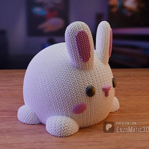 May include: A white knitted bunny plush toy with pink ears and a pink nose. The bunny has black eyes and a white and pink knitted texture.