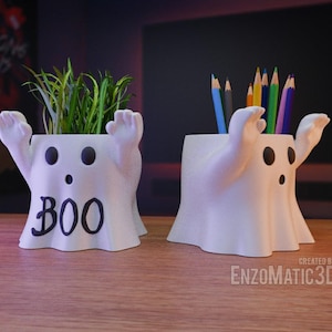 May include: Two white ghost-shaped pencil holders. One holds a bunch of colored pencils and the other holds a small potted plant. Both have a friendly face with the word "BOO" written on the front.