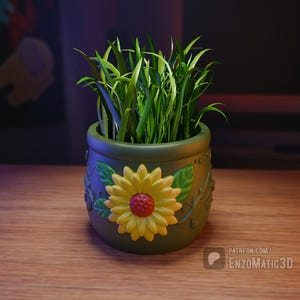 Può includere: Un vaso di ceramica verde con un disegno di girasole e foglie verdi. Il vaso è pieno di erba verde.
