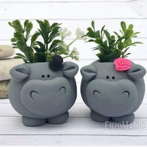Puede incluir: Dos macetas de cerámica gris en forma de hipopótamo con plantas verdes y una flor rosa. El hipopótamo de la izquierda lleva un sombrero de copa negro.