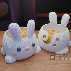 May include: Two white ceramic yarn bowls shaped like bunnies. The bowls have pink ears and a knitted texture. One bowl has a ball of yarn inside.
