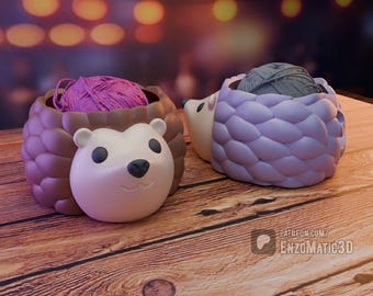 May include: Two hedgehog-shaped yarn bowls. One is brown with a pink yarn ball, and the other is lavender with a gray yarn ball. The bowls have a cute face design and are sitting on a wooden surface.
