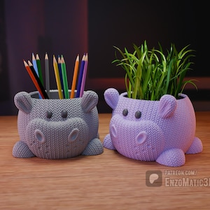 May include: Two hippo-shaped planters, one gray and one lavender, sit side-by-side on a wooden surface. The gray planter holds colored pencils, while the lavender one contains green plants. Both have a knitted texture and cute details.