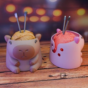 May include: Two whimsical yarn holders, one beige and one pink, each shaped like a cartoon animal. The beige holder is a sheep, and the pink one is a pig. Each holds a ball of yarn and a pair of knitting needles. The yarn is yellow and pink.