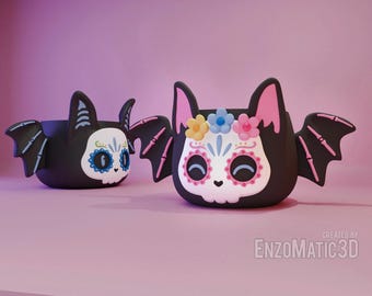 May include: Two black bat-shaped planters with white sugar skull faces and colorful floral details. The planter on the left has blue eyes and the planter on the right has pink eyes.