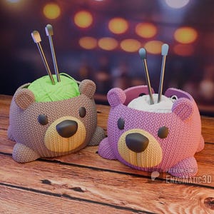 Puede incluir: Dos sujetahilos con forma de oso, uno marrón y otro morado, sobre una superficie de madera. Cada oso tiene una nariz grande y ovalada y contiene hilo y agujas de tejer. El oso marrón contiene hilo verde, mientras que el oso morado contiene hilo blanco.