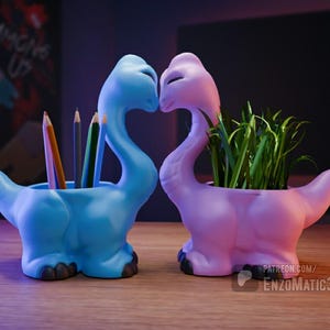 Puede incluir: Dos macetas con forma de dinosaurio, una azul claro y otra lavanda, están una al lado de la otra. El dinosaurio azul contiene lápices, mientras que el lavanda contiene plantas verdes. Las macetas están sobre una superficie de madera.