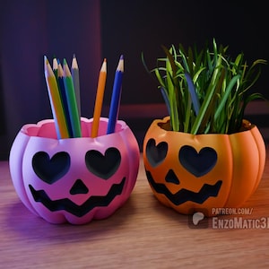 Puede incluir: Dos porta lápices de color naranja y rosa con forma de calabaza con ojos en forma de corazón y una boca sonriente. El porta lápices rosa tiene lápices de colores dentro y el porta lápices naranja tiene hierba verde dentro.