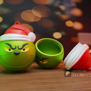May include: A green, spherical ornament featuring the Grinch's face, complete with a Santa hat. The ornament is open, revealing a hollow interior. A red and white ornament is also present. The text "PATREON.COM/EnzoMatic3D" is visible.