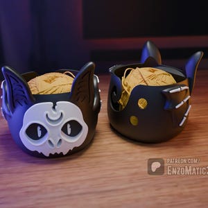 May include: Two black yarn bowl cat head designs with a white face and a crescent moon on the forehead. One cat head is facing the left and the other is facing the right. Both bowls are filled with yarn.