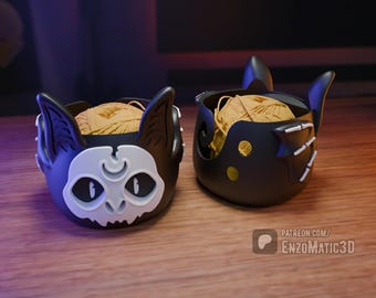 May include: Two black yarn bowl cat head designs with a white face and a crescent moon on the forehead. One cat head is facing the left and the other is facing the right. Both bowls are filled with yarn.