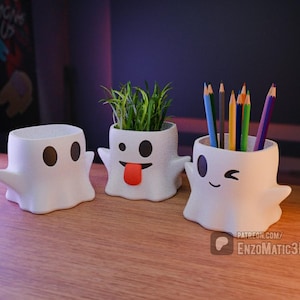 May include: Three white ghost-shaped ceramic planters with different facial expressions. The planter on the left has two eyes, the middle planter has a tongue sticking out, and the planter on the right has a winking eye. The middle planter has a small green plant inside.