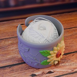 May include: A lavender yarn bowl with a white ball of yarn inside. The bowl has a sunflower design on the side and a floral vine pattern. The bowl is sitting on a wooden surface.