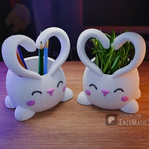 May include: Two white ceramic bunny-shaped planters with pink accents. One planter has green grass growing inside, and the other has colorful pencils inside.
