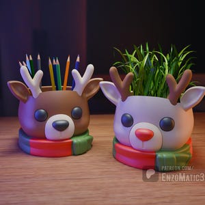 Puede incluir: Dos recipientes con forma de reno, uno marrón y otro blanco, cada uno con una bufanda roja, verde y blanca. El reno marrón contiene lápices de colores, mientras que el blanco contiene plantas verdes. Sobre una superficie de madera.