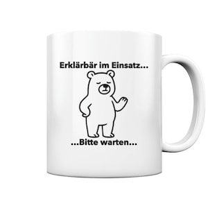 Peut inclure: Mug en céramique blanche avec un ours noir et blanc. Le texte sur la tasse est "Erklärbär im Einsatz..." et "...Bitte warten...". L'ours est dessiné dans un style simple et amical.