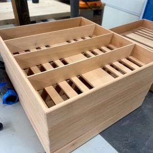 May include: A light-colored wooden storage tray featuring a slatted base and three internal dividers that create four distinct rectangular compartments. The tray has smooth, straight sides and an open top, constructed from natural light wood.