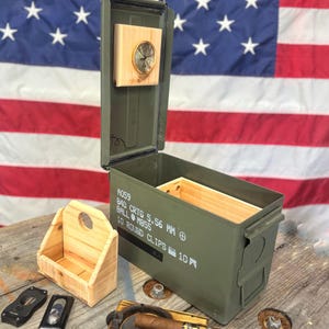 以下が含まれることがあります： オリーブグリーンの弾薬箱。内部は木製で、蓋の内側には小さな時計が取り付けられています。葉巻用の木製レスト、葉巻、小さな木箱、2つのライターも写っています。背景にはアメリカ国旗があります。