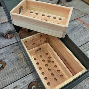 May include: Two rectangular light-colored wooden trays with perforated bases and circular side cutouts, designed to fit inside an olive green metal ammunition box. The trays feature visible wood grain and are stacked within the container.