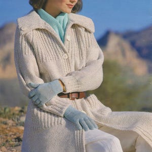Op de afbeelding: Een vrouw draagt een lange, witte gebreide cardigan met een lichtblauwe sjaal en bijpassende handschoenen. Ze zit op een rots in een woestijnlandschap.