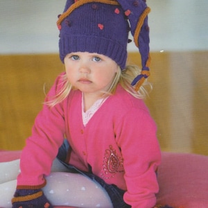 May include: A child wearing a purple knit hat with red heart accents and a long, pointed end. The hat has orange trim and is paired with a pink cardigan and matching knit booties. The outfit is on a pink surface.