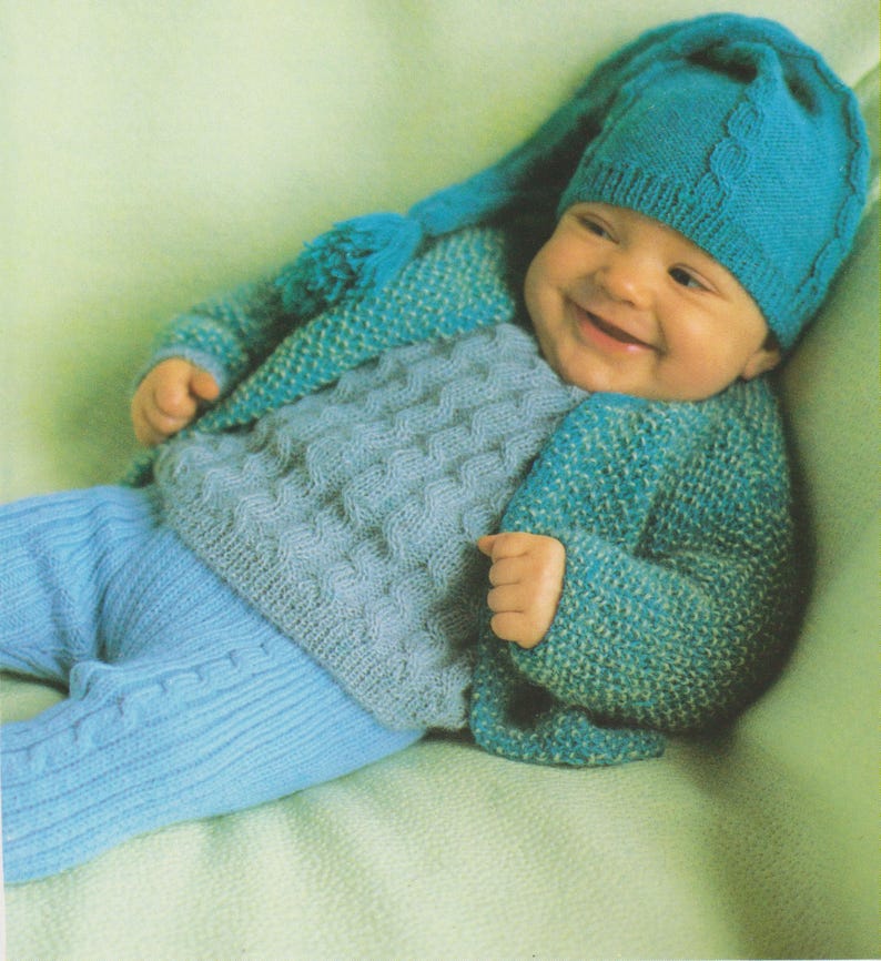 Puede incluir: Un beb&eacute; con un gorro azul tejido con una punta larga, un su&eacute;ter azul tejido con un su&eacute;ter azul claro tejido debajo y pantalones azules claros tejidos.