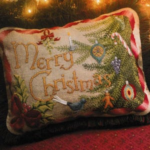 Puede incluir: Cojín rectangular bordado con temática navideña. Las palabras "Merry Christmas" están bordadas con hilo dorado. También se borda un árbol de Navidad, adornos y un bastón de caramelo. El cojín tiene un borde rojo y está sobre una superficie roja.