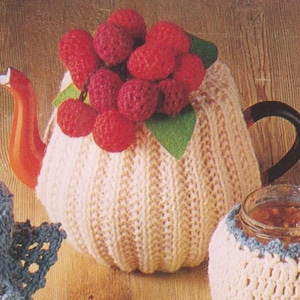 May include: A cream-colored knitted teapot cozy adorned with a cluster of red knitted berries and green leaves. The teapot has an orange spout and a black handle. A jar of jam is also visible.