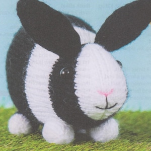 May include: A black and white knitted rabbit toy with black ears and a pink nose. The rabbit is sitting on a green grassy surface.